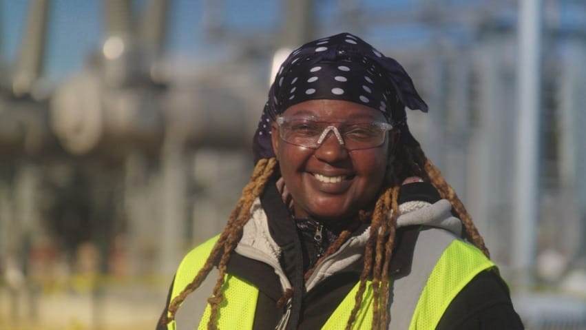 Worker in safety gear and yellow vest smiles at an industrial site with machinery in the background.
