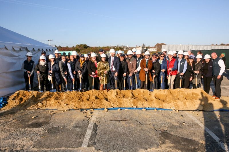 Group of people in hard hats participating in a groundbreaking ceremony with shovels and a pile of dirt in front of a tent