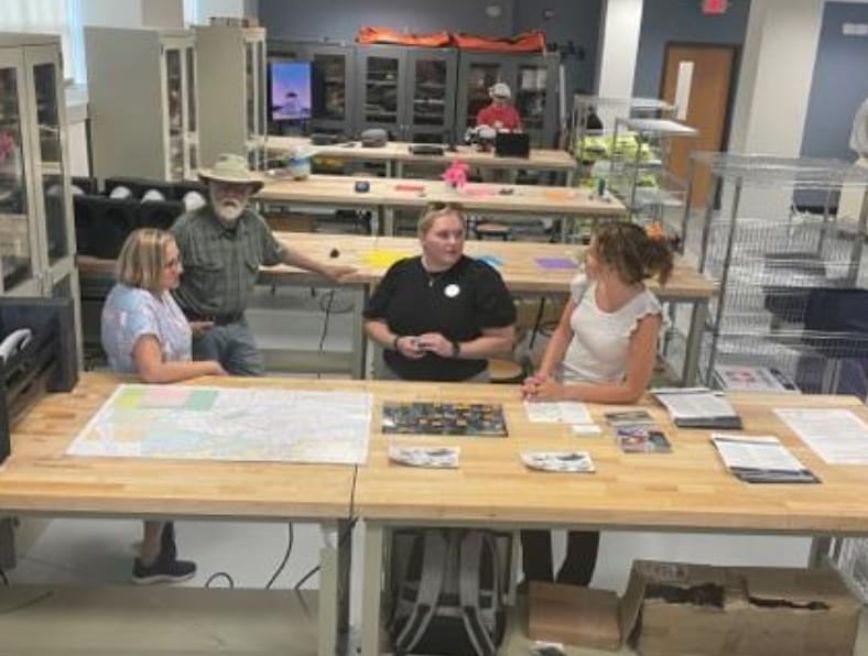Four people having a discussion around a large table with maps and documents in a room with shelves and equipment.
