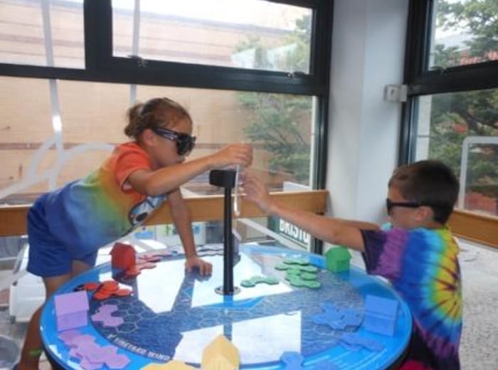 Children playing a tabletop interactive game with colored shapes and wearing sunglasses indoors.