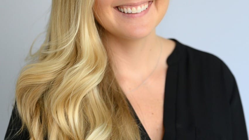 Smiling person with long blonde hair wearing a black top against a plain background.