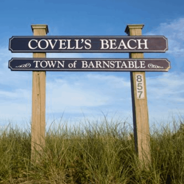 Sign for Covell's Beach, Town of Barnstable, with tall grass and a clear blue sky background.