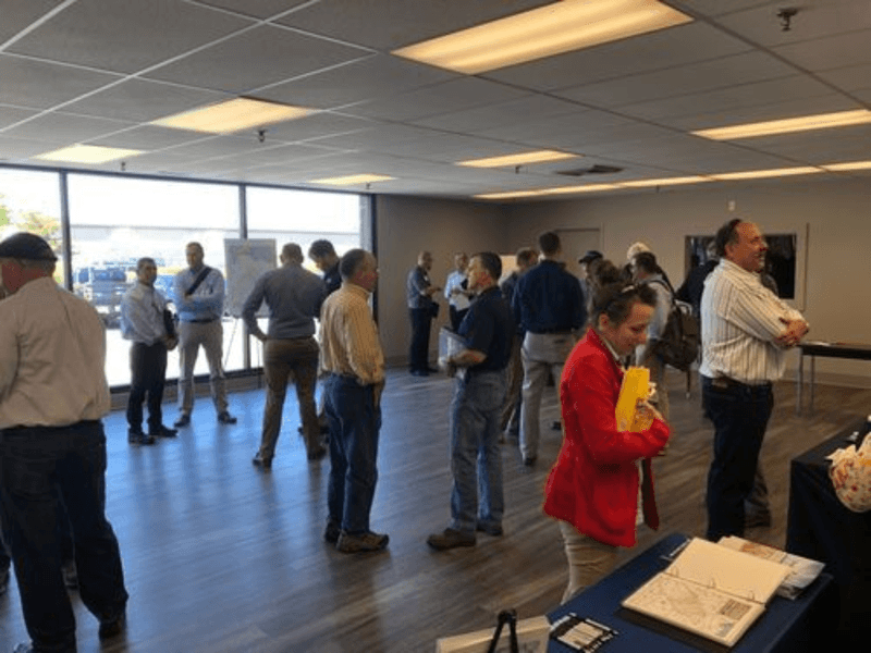 People standing and chatting in a large room with tables and natural lighting.