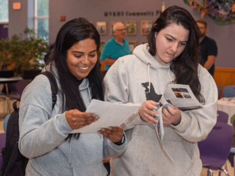 Two people in hoodies reading documents in a community center setting with others in the background.