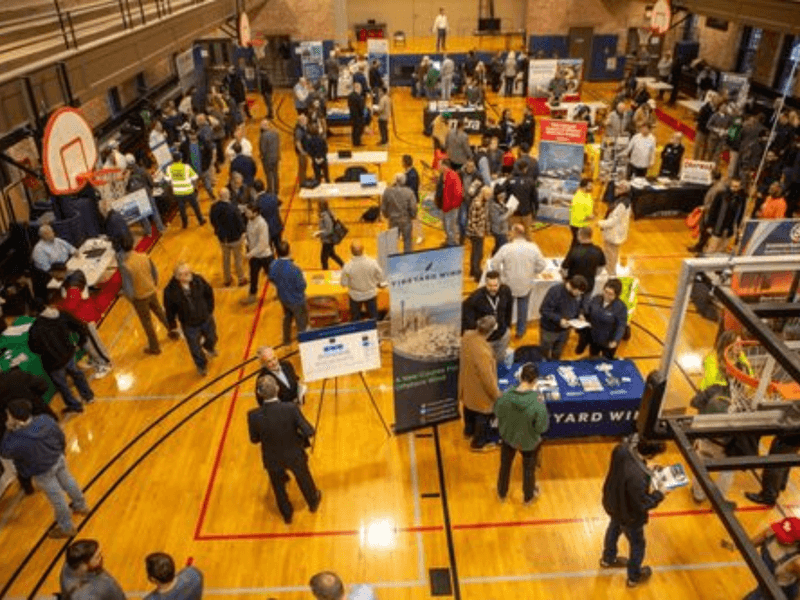 Career fair in a gymnasium with people visiting booths and engaging with company representatives.