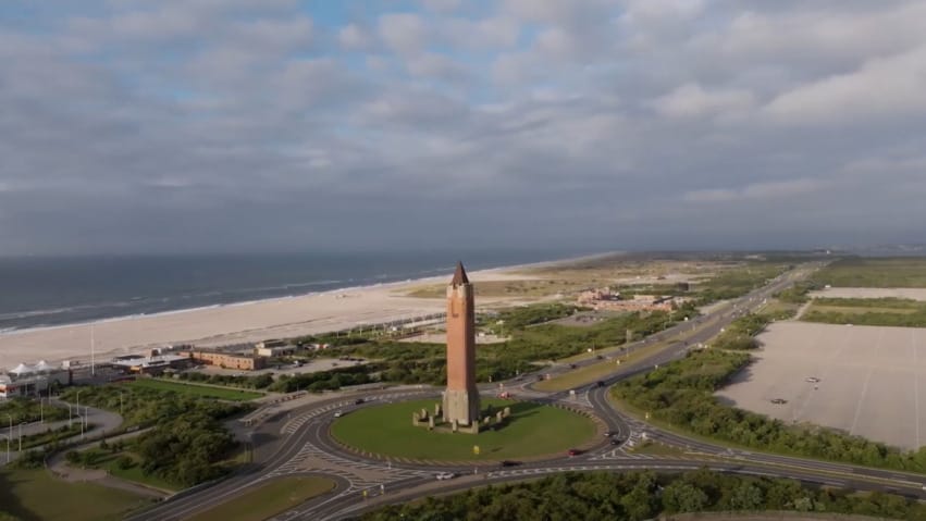 Aerial view of a tall tower surrounded by a circular road, near a coastline with a sandy beach and blue sky.