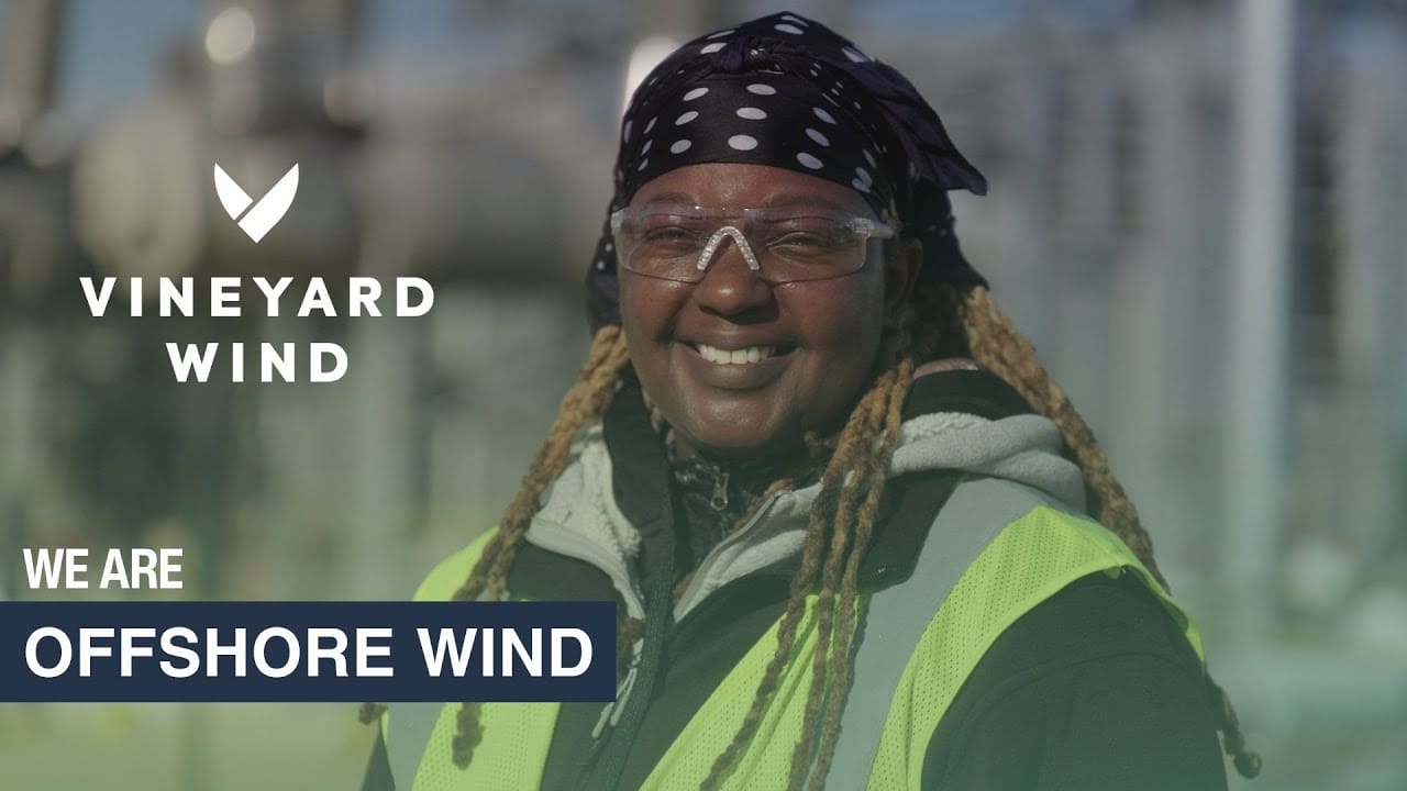 Person in safety gear smiling at an offshore wind site, Vineyard Wind logo visible.