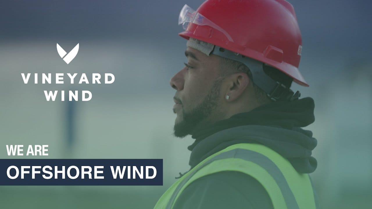 Worker in red hard hat and safety vest looking at Vineyard Wind turbine construction site.