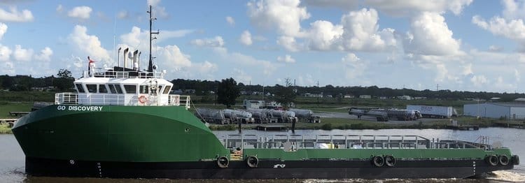 Green and white vessel named "Go Discovery" on a river with a background of industrial buildings and partly cloudy sky.