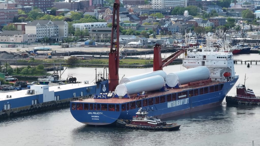 Cargo ship with wind turbine components docked in a harbor, surrounded by buildings and small tugboat nearby.