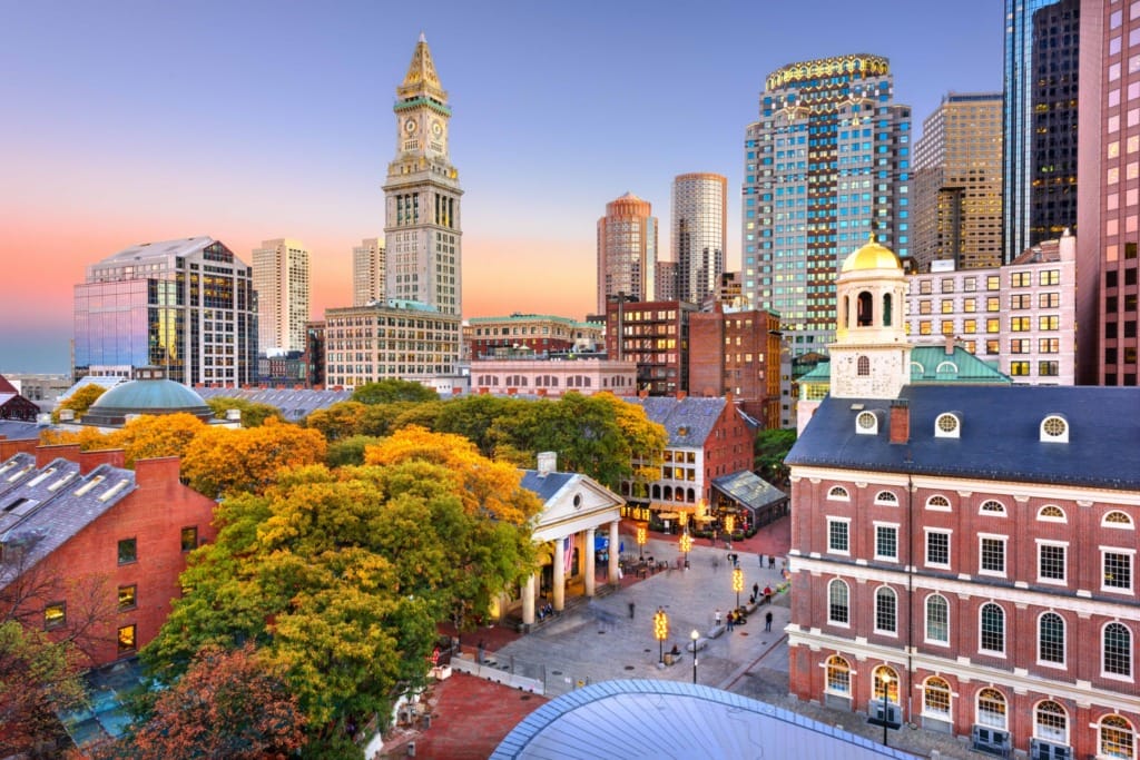 Boston cityscape at sunset with historical buildings, modern skyscrapers, and autumn trees in the foreground.