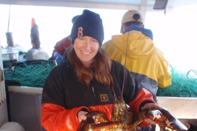 Person holding a large lobster on a fishing boat, wearing a beanie and waterproof clothing, with nets in the background.