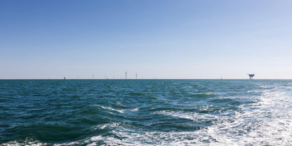 Wide ocean view with distant offshore wind turbines under a clear blue sky, waves gently rolling in.