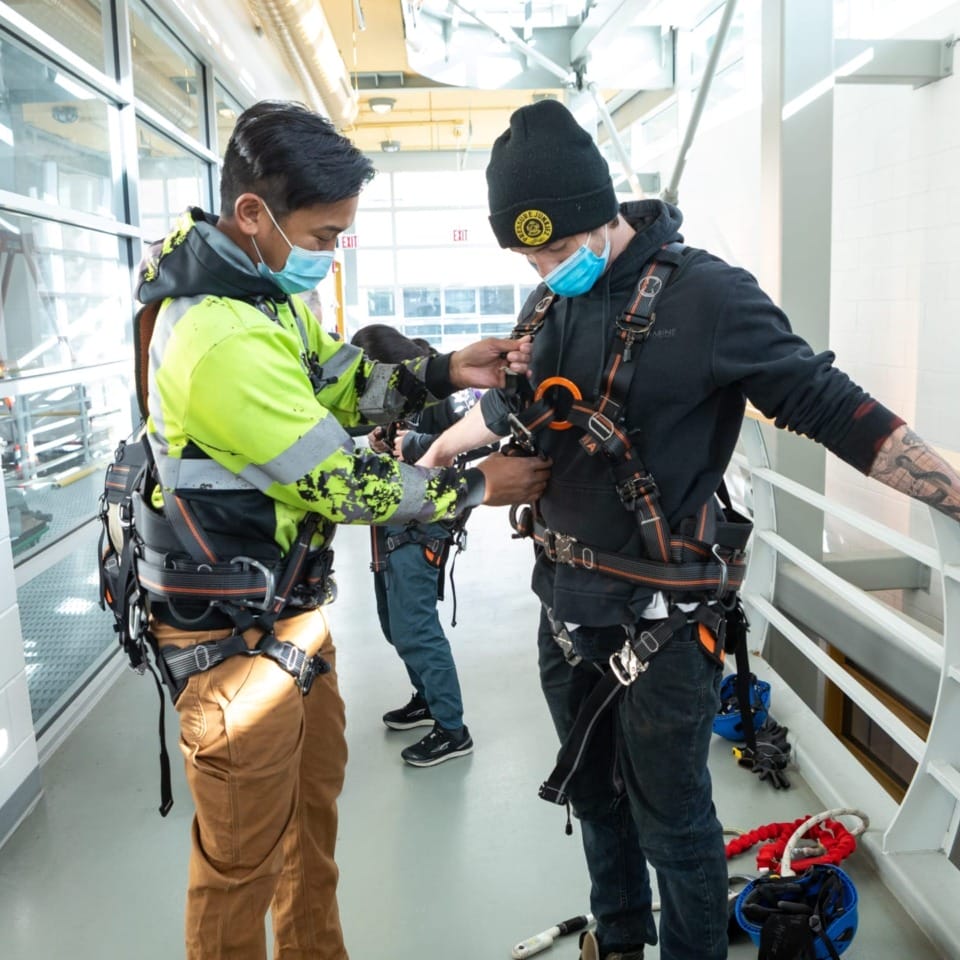 Two workers in safety harnesses adjusting gear indoors wearing masks.