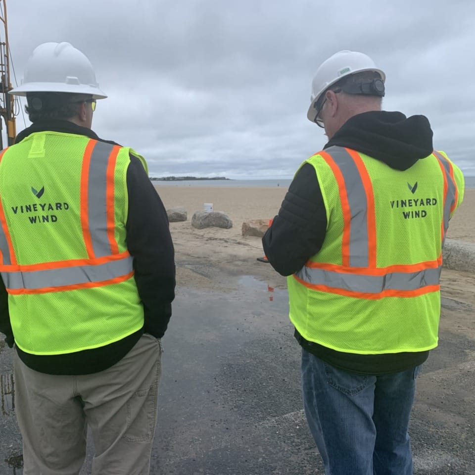 Two workers in safety vests and helmets at a construction site near a beach, with overcast skies in the background.