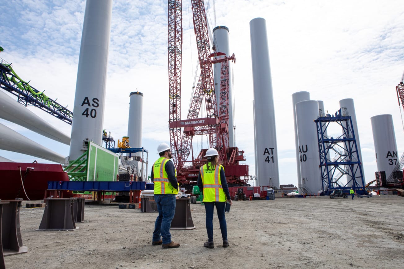 Workers in safety vests at a construction site with large cranes and tall cylindrical structures in the background.