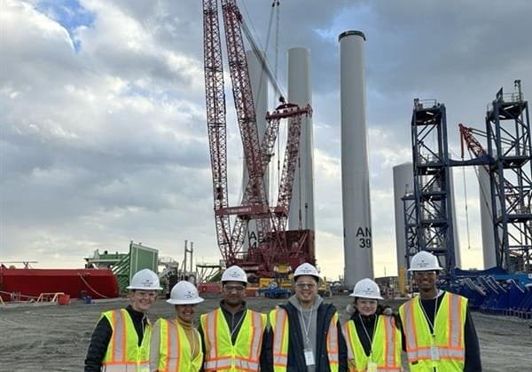 Construction crew in safety gear standing at a construction site with large cranes and towers in the background.