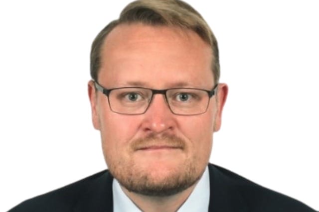 Man with glasses wearing a suit and tie, looking directly at the camera, white background.