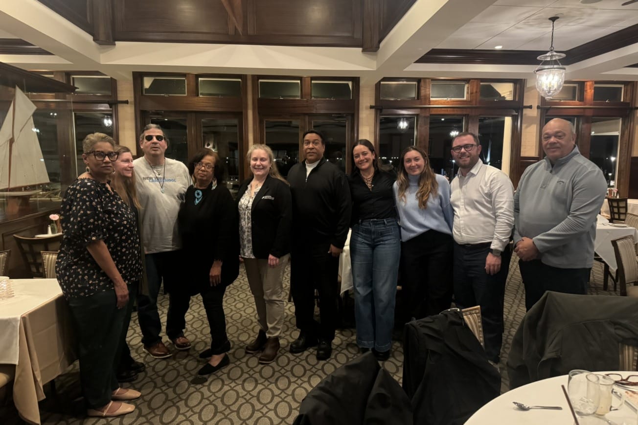Group of people smiling and posing indoors at a gathering event in a warmly lit room with large windows and wooden accents