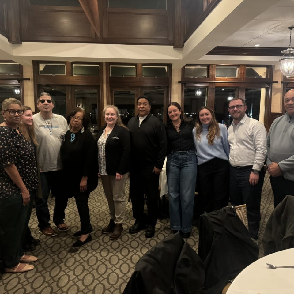 Group of people smiling and posing indoors at a gathering event in a warmly lit room with large windows and wooden accents