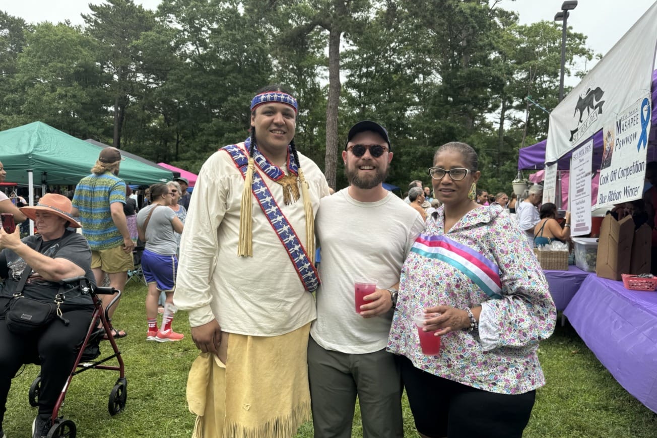 Three people posing together at an outdoor event with tents and trees in the background