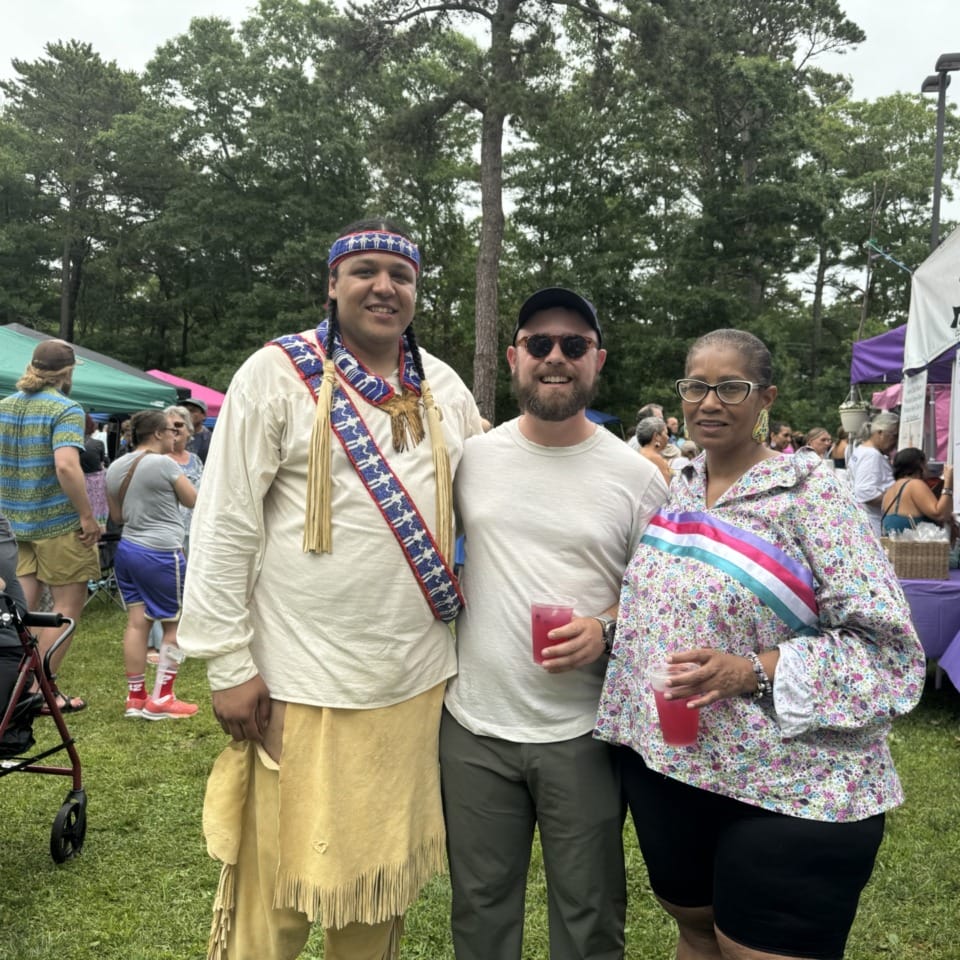 Three people posing together at an outdoor event with tents and trees in the background