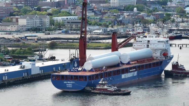 Cargo ship unloading large cylindrical components at a bustling harbor with a tugboat assisting nearby.