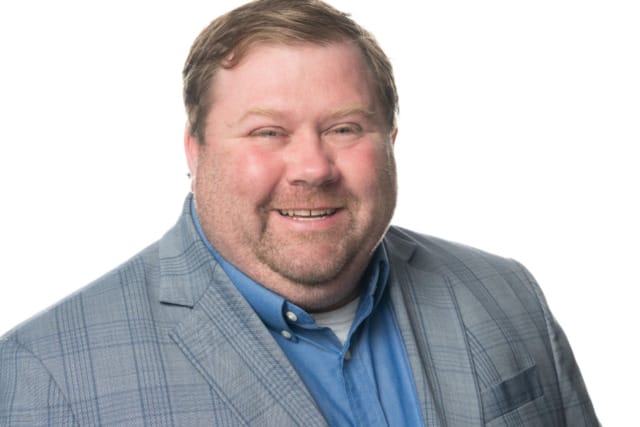 Man in a blue shirt and plaid suit jacket smiling against a white background.