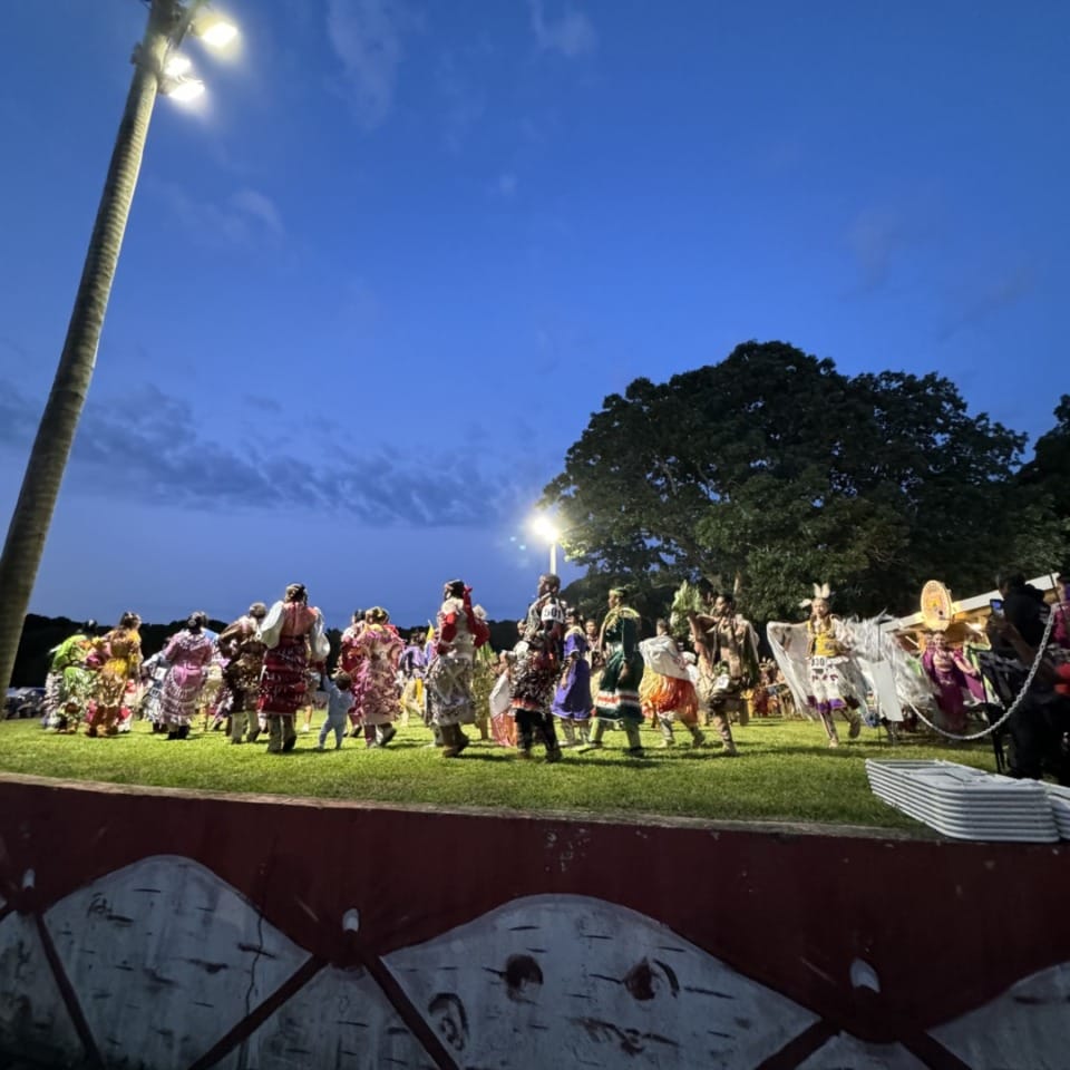 Colorful cultural performance taking place outdoors at dusk with a group of people in traditional attire, stage decorated.