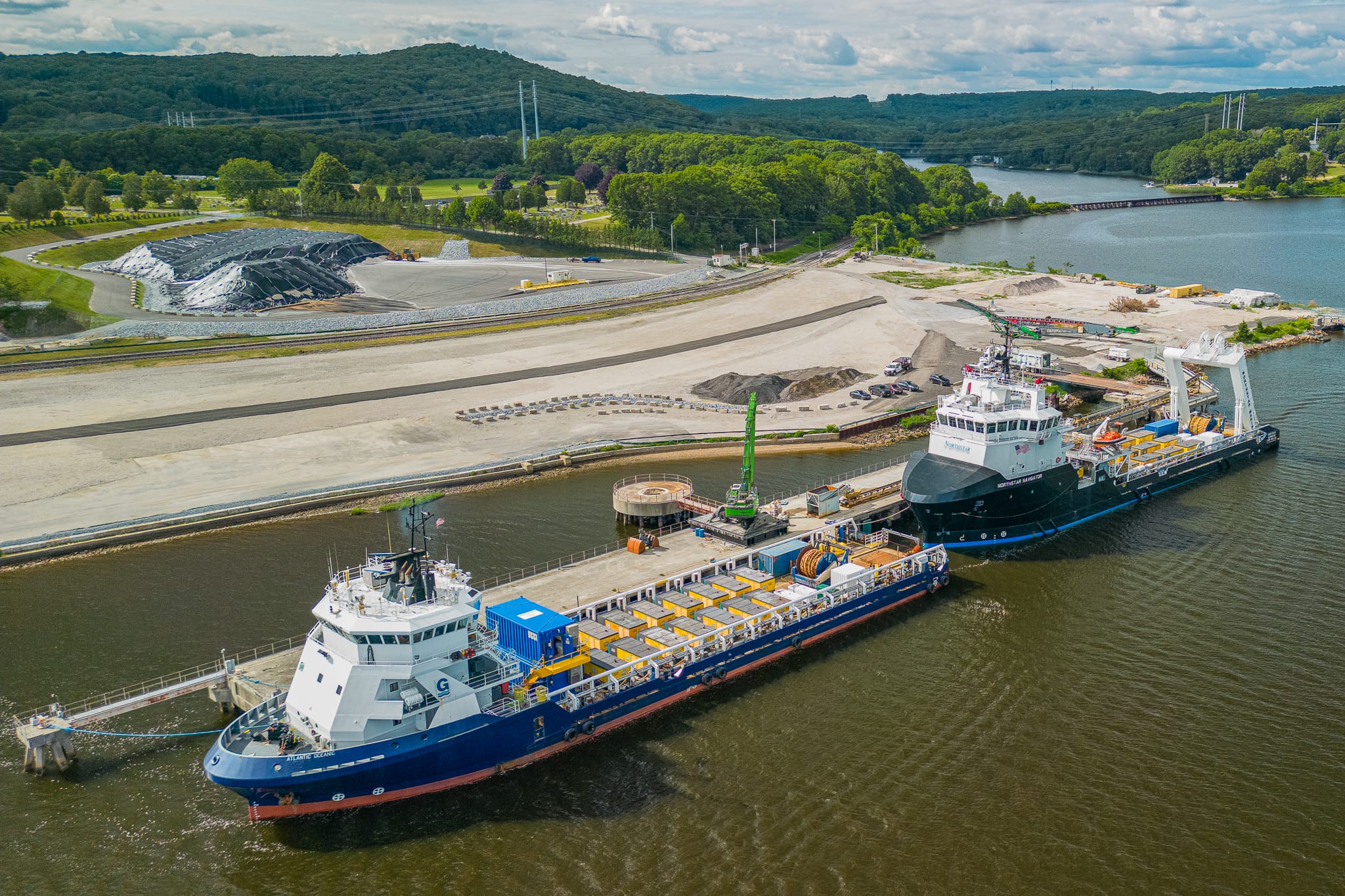 Two cargo ships docked at an industrial port with lush green hills and a cloudy sky in the background.