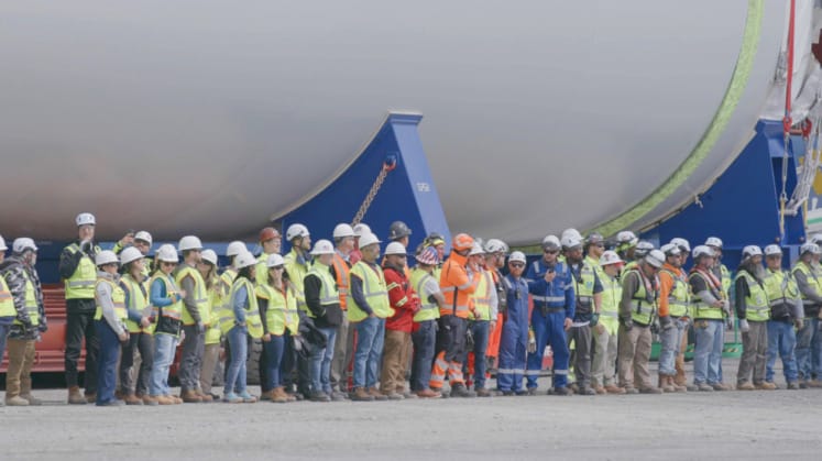Construction workers in safety gear standing in line near a large industrial tank on a construction site.