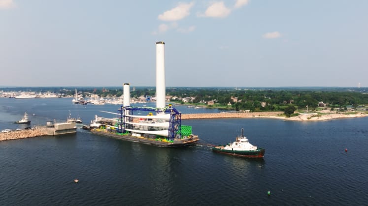 Aerial view of a large ship with two tall structures being guided by a tugboat in a coastal harbor with a city in the background