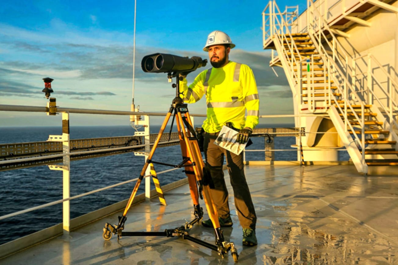 Worker on offshore platform using a large binocular telescope for observation, wearing high-visibility gear and helmet.
